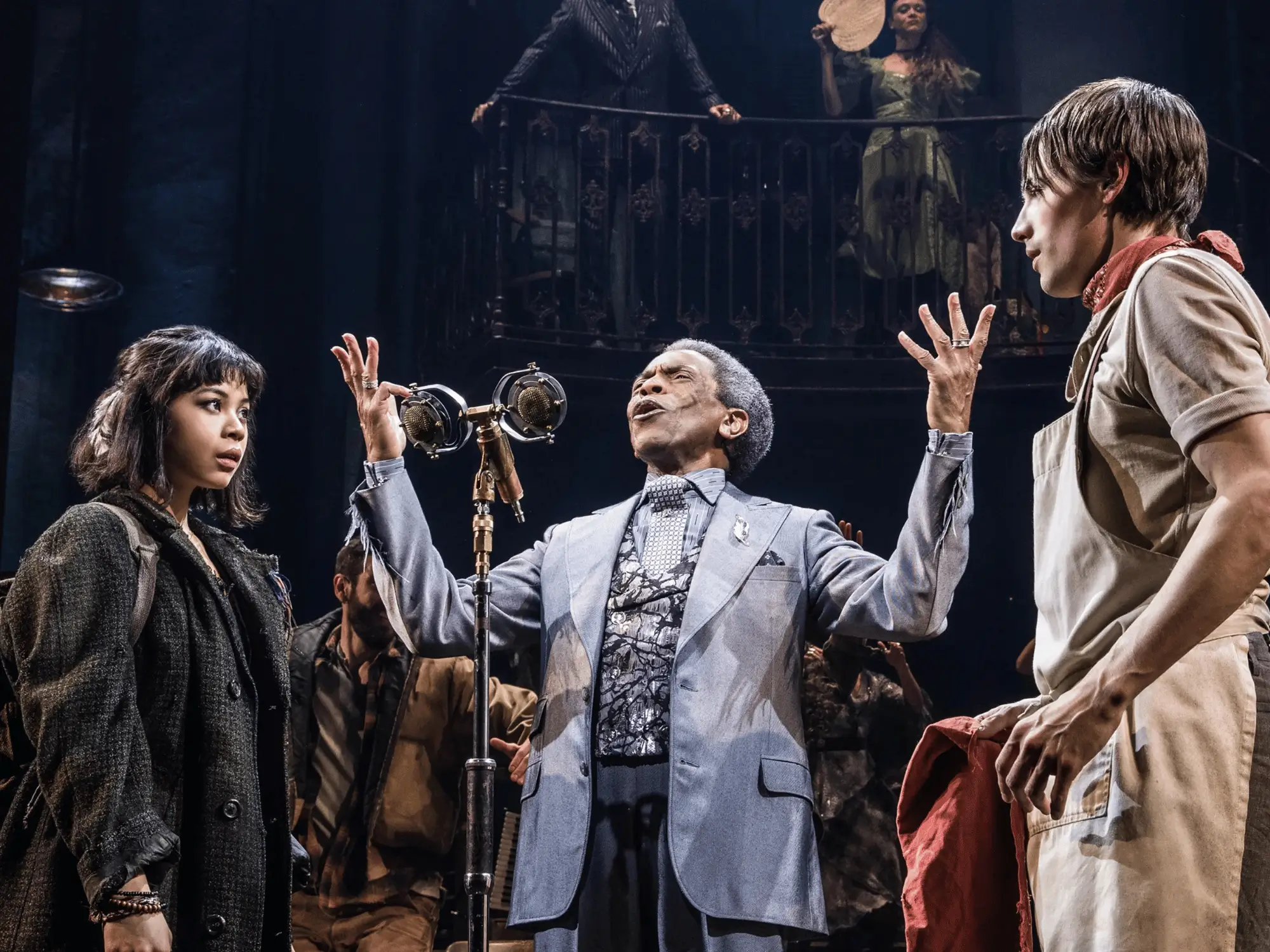 Three performers gathered around a vintage silver microphone in a scene from Hadestown