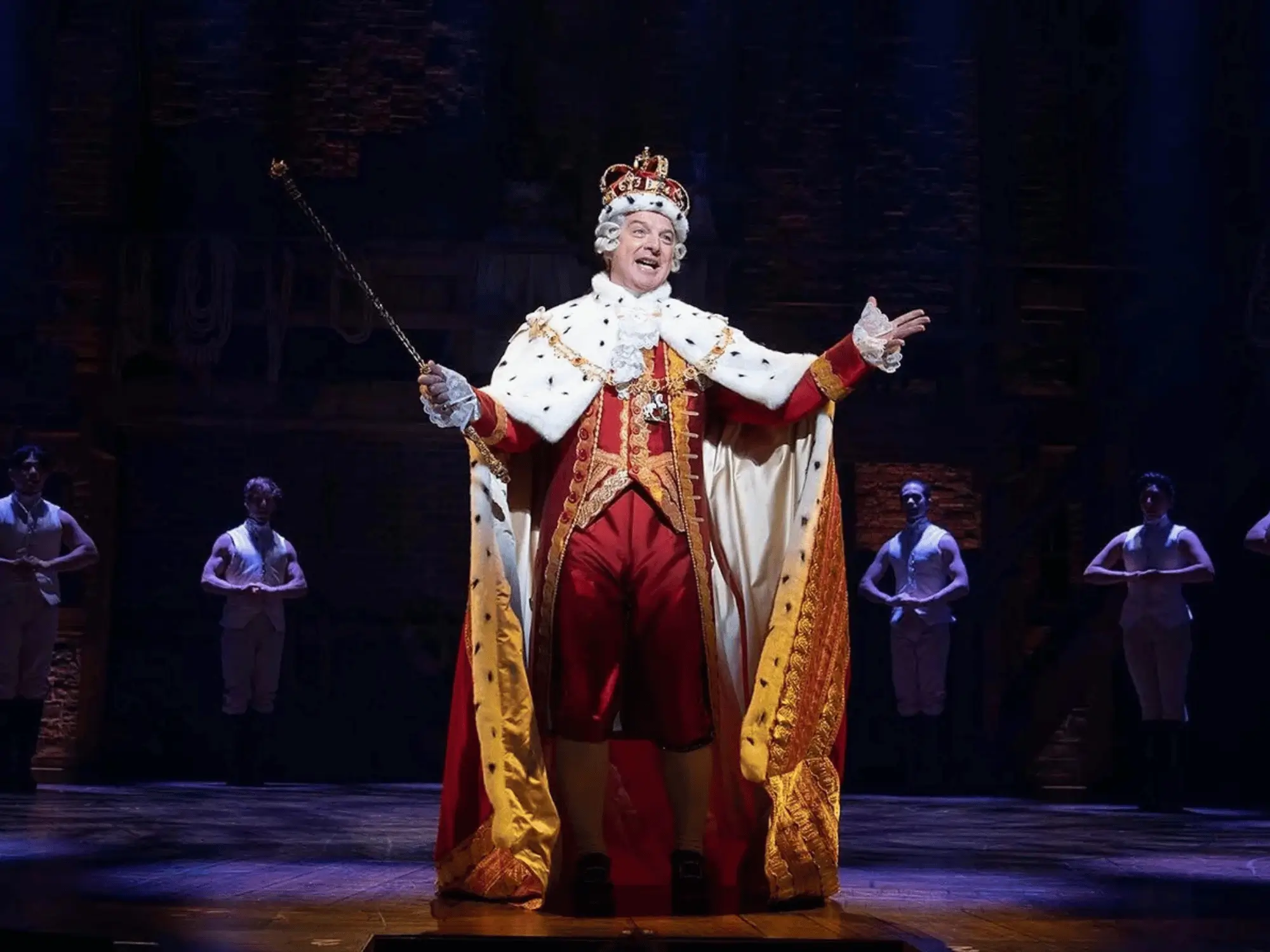 A performer in a regal crown and jeweled robes singing center stage in a Broadway musical