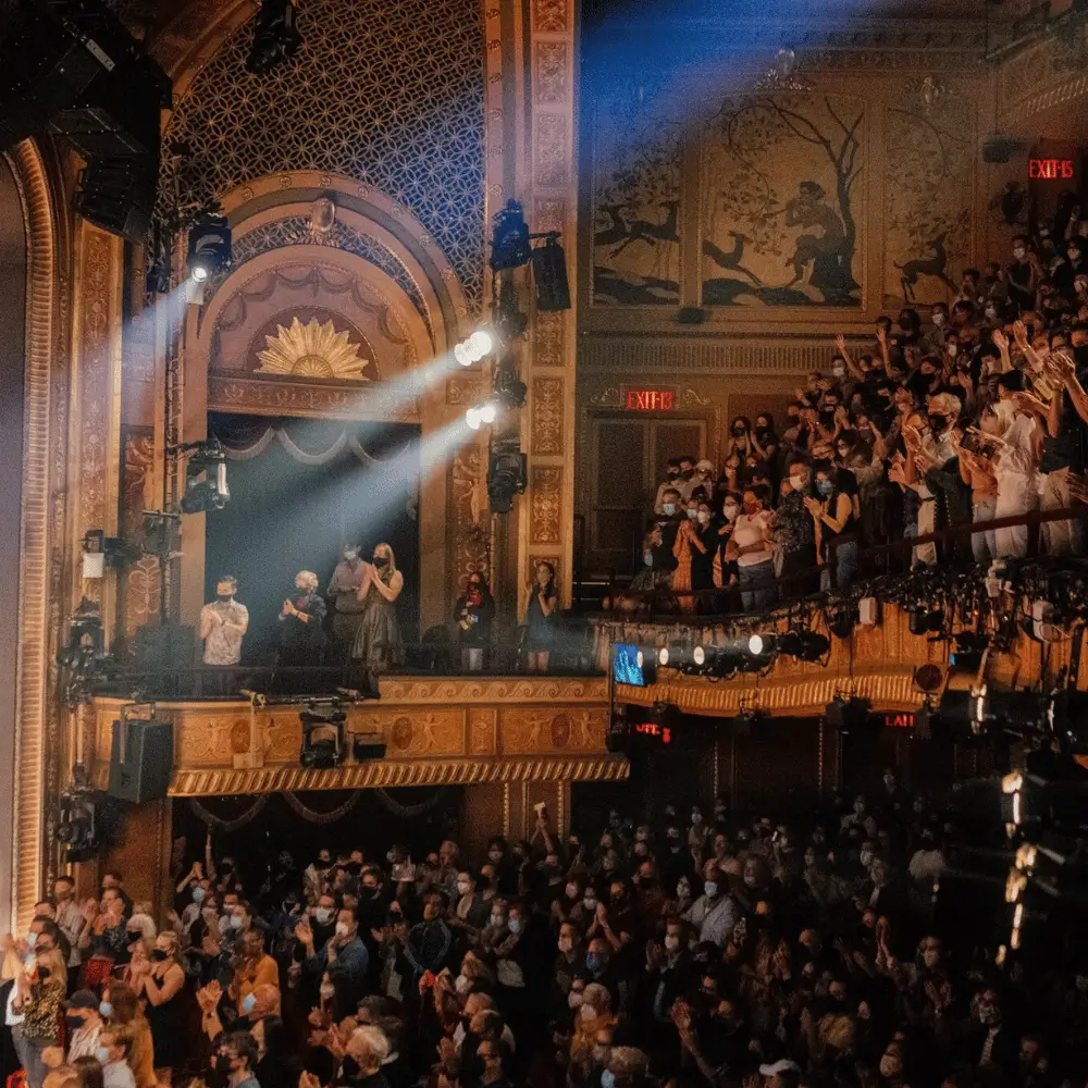 A packed theater audience applauding from the orchestra and balcony levels