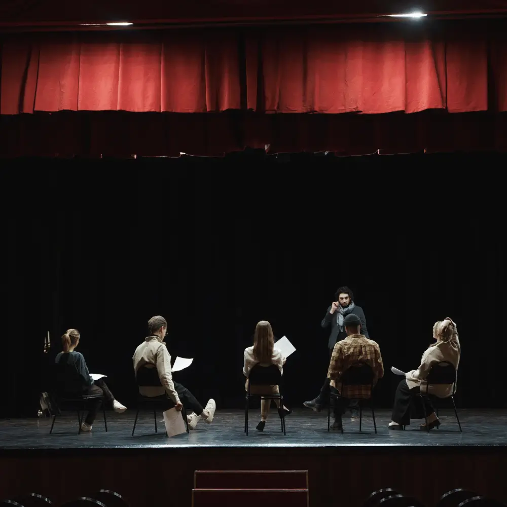 A director leading a script reading with actors seated in a circle on stage